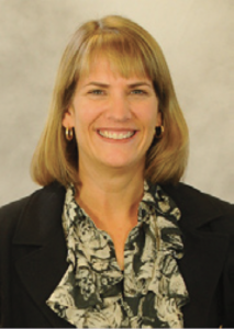 Denise Daniels with shoulder-length blonde hair, black blazer, patterned blouse, seated indoors in an academic setting.